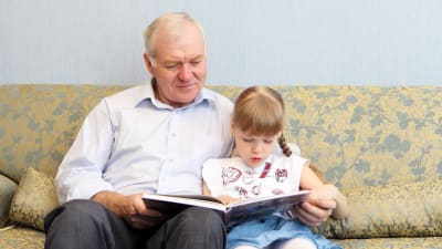 Äldre man och flicka läser en bok tillsammans i soffan. 