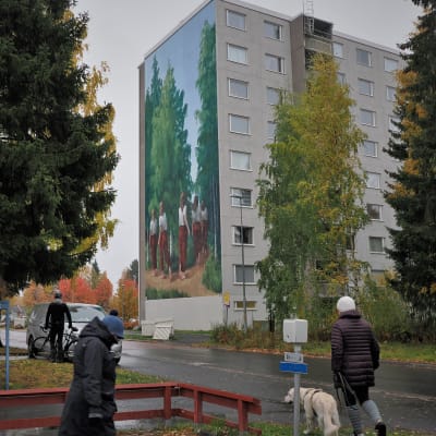 Ihmisiä kävelee etualalla ja taustalla kerrotalo, jonka seinään on maalattu isokokoinen muraali. 
