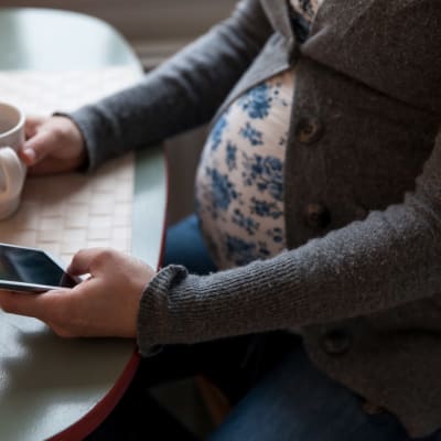Gravid kvinna sitter och dricker kaffe och använder sin mobiltelefon.