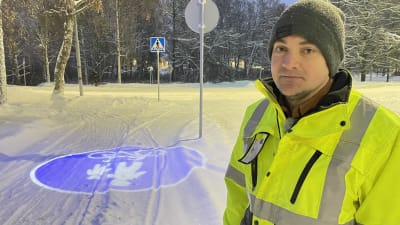 En man i gul reflexjacka och mössa står på en snöig cykelväg med ett upplyst trafikmärke på marken. 