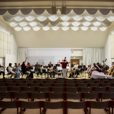 Orkesteri harjoittelee konserttisalin lavalla, etualalla näkyy katsomon tyhjiä penkkejä.