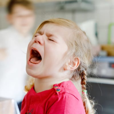 En liten upprörd flicka sitter i köket och skriker med öppen mun och slutna ögon.