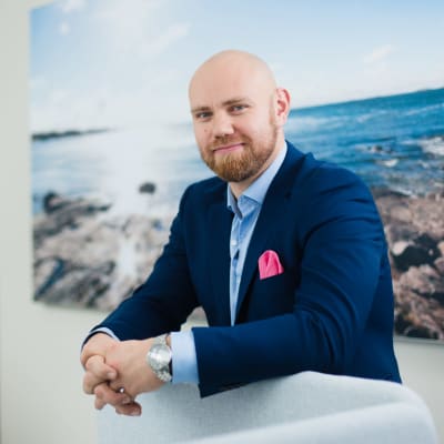 Aki Eriksson klädd i mörkblå kavaj står i ett kontor framför en stor väggplansch med klippor och hav.