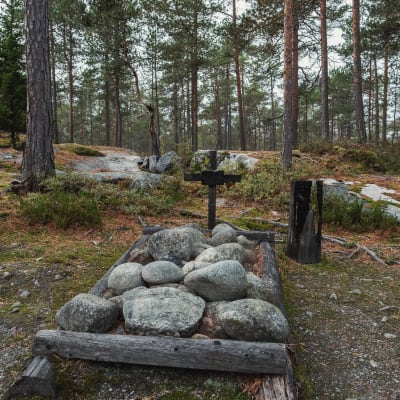 Kiviä maassa aseteltuna puisen kehikon sisään, risti, taustalla metsäistä maisemaa, pohjoisgrönlantilainen hauta