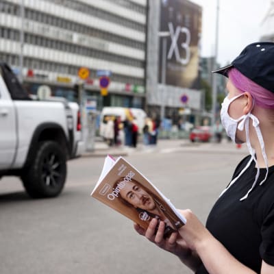 Nuori nainen lukee opinto-opasta kasvomaski naamallaan Helsingin keskustassa.