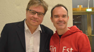Pekka Sauri och Peter Vesterbacka står bredvid varandra i halvfigur, bilden är tagen i vestibulen på Helsingfors stadshus. Sauri har kavaj och vit skjorta utan slips, Vesterbacka röd luvtröja med texten FinEst.