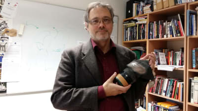 Professor Mats FRidlund, med gasmask  handen, framför sin bokhylla