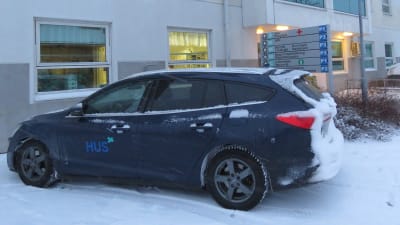 En blå personbil med HUS logga på dörren. Det är vinter, bilen som har lite snö på sig står parkerad invid en byggnad, Raseborgs sjukhus