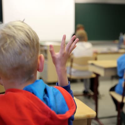 En blond elev fotograferad bakifrån med röd tröja markerar i en skolklass. 