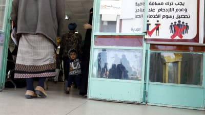 Ett barn står i dörren till en läkarmottagning i Jemen. Bredvid dörren finns en skylt som uppmanar besökarna att undvika folksamlingar