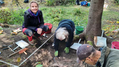 Två personer gräver i en grop, en tredje sitter på huk vid kanten av gropen. Det är en kvinna som tittar in i kameran och ler.
