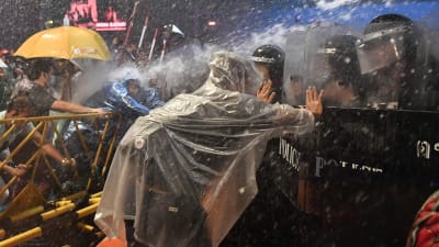 Person iförd i regnrock som trycker mot polisens kravallsköld.