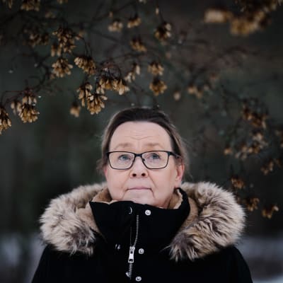 Päivi Grönlund seisoo lehtensä pudottaneen puun alla.