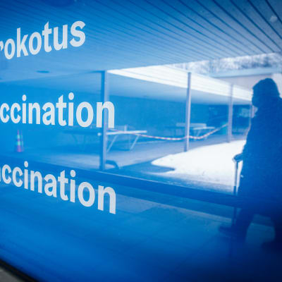 Skylt där det står Coronavaccination. I reflektionen av skylten syns en person som promenerar.
