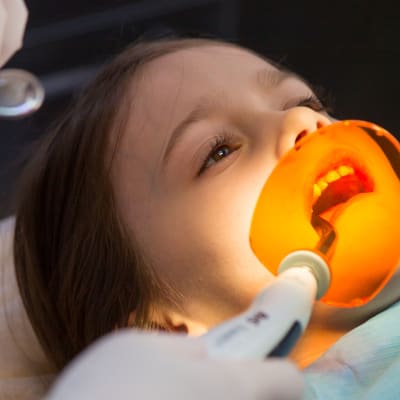 En flicka ligger i tandläkarstolen med tandläkarinstrument i munnen.