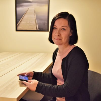 Pia Köpsi, en dam med rosa skjorta och svart tröja, sitter vid ett skrivbord.