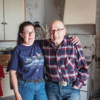 Kvinna med mörkt hår i ponnysvans, glasögon, t-skjorta och jeans håller armen om axlarna på äldre man klädd i rutig flanellskjorta och jeans.