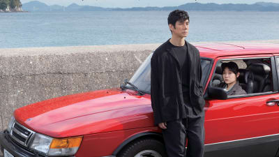 Mustiin pukeutunut mies nojaa punaiseen Saabiin, nuori naiskuljettaja katselee ulos, kuva elokuvasta Drive My Car.