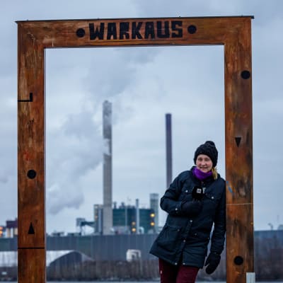 Yle Varkauden toimittaja Tarja Nyyssönen seisoo suuren taulukehyksen sisällä. Taustalla järven takana näkyy Stora Enson tehdas.