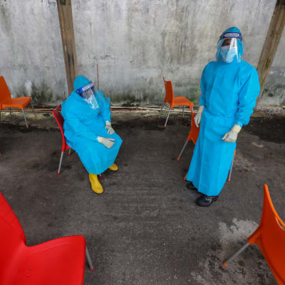 Två personer klädda i blåa skyddsoveraller, handksar och ansiktsskydd tar en paus. runt omkring dem står färggranna plaststolar.