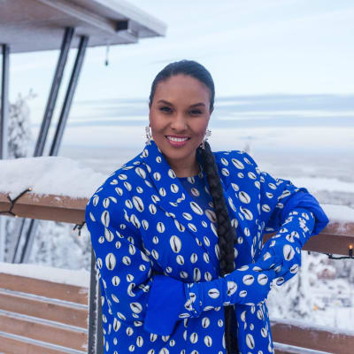 Värikkääseen asuun pukeutunut pitkähiuksinen nainen katsoo kameraan hymyillen. Hän nojaa kausivalojen koristamaan kaiteeseen ja hänen takanaan avautuu luminen alppikylämaisema.