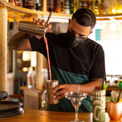 En bartender med munskydd på häller en drink i ett glas.