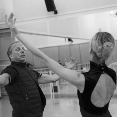 Mustavalkoisessa kuvassa keski-ikäinen mies ohjaa selin kameraan olevaa balettitanssijaa harjoitussalilla, kummallakin kädet levitettyinä.