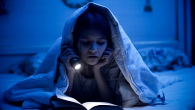 En liten flicka läser med ficklampa under täcket i ett mörkt rum.