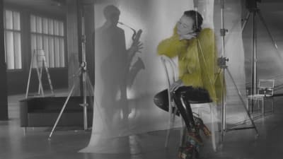 Runoilija Elsa Tölli istuu teollisuushallissa yllään keltainen turkispuuhka. Hän nojaa päätään kättään vasten. Hänen takanaan olevalla kankaalla näkyy saksofonisti Linda Fredrikssonin varjo.