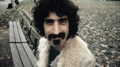 Frank Zappa hymyilee puolilähikuvassa puiston penkillä.