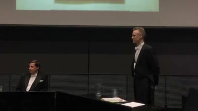 Två män klädda i frack i ett auditorium framför en presentationsskärm.