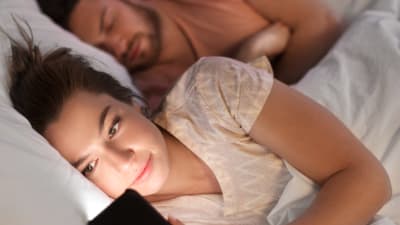 Kvinna ligger och tittar på telefonen medan mannen bakom henne sover. 