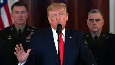 Donald Trump talar vid ett podium, två män i militärunifrom står bakom honom. 