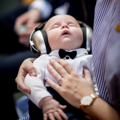 En sovande bebis i en vuxen människas famn. Bebisen har stora hörlurar som skyddar hörseln.
