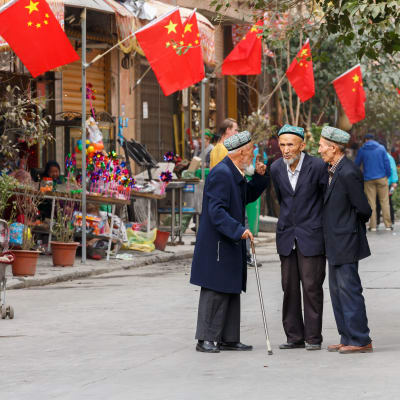 Tre äldre män från den uiguriska minoriteten  på en gata i den gamla oasstaden Kashgar i provinsen Xinjiang i det västligaste Kina.  Kinesiska flaggor i bakgrunden.