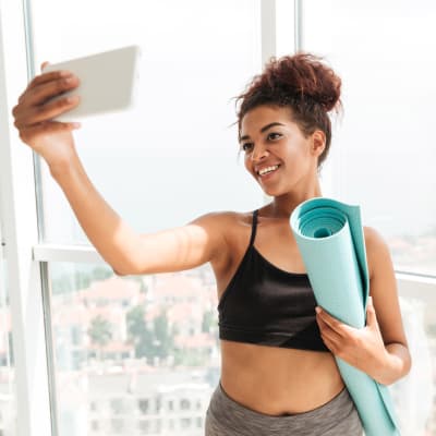 Leende ung kvinna med hoprullad yogamatta under armen tar en selfie