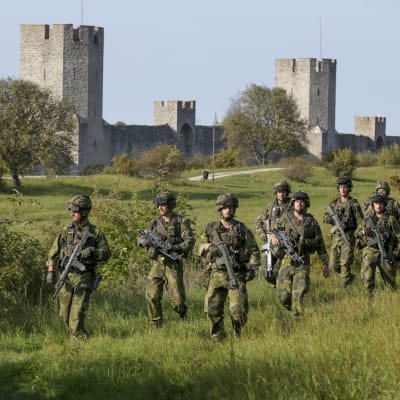 11 militärer går på en äng i full mundering och vapen i hand. I bakgrunden syns Visby ringmur.