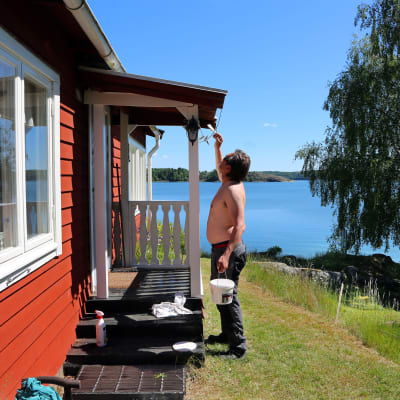 Person målar röd stuga med vita knutar vid strand. 