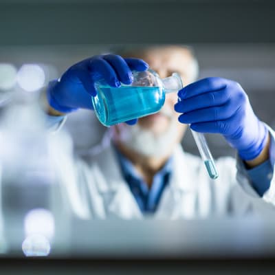 En manlig forskare med skägg och blåa handskar  häller blå vätska i ett provrör i ett laboratorium. Ansiktet syns inte. Snygg bildsättningsbild.