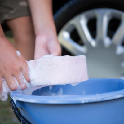 Händer som sköljer tvättsvamp i fat vid ett bilhjul.