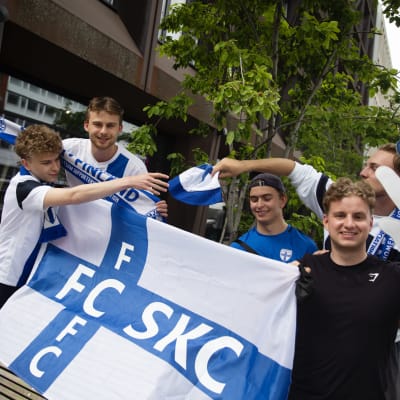 Fem glada unga män håller upp finska flaggor och fotbollshalsdukar