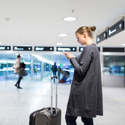 En kvinna står på ett flygfält och tittar på sin mobil, med kappsäcken framför sig.
