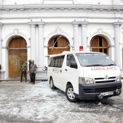 En ambulans står parkerad utanför ett hotell som utsatts för en explosion. Flera beväpnade poliser syns på bilden och marken är täckt av glassplitter.
