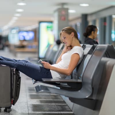 Kvinna sitter tillbakalutad på bänk i en terminal. Hon har fötterna på sin resväska och surfar på sin pekplatta.