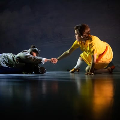 En kvinna i gul klänning sitter på huk på en teaterscen och drar upp en kvinna i grön jacka ur en isvak