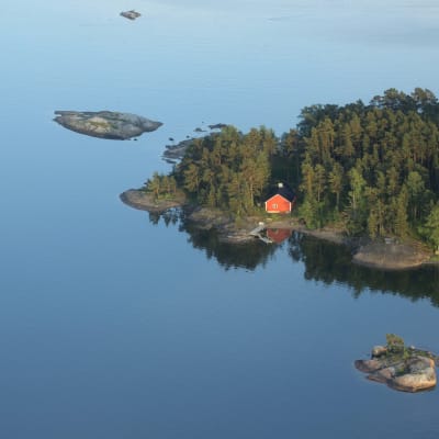 En ö med en röd stuga och blått vatten omkring i Ingå skärgård.