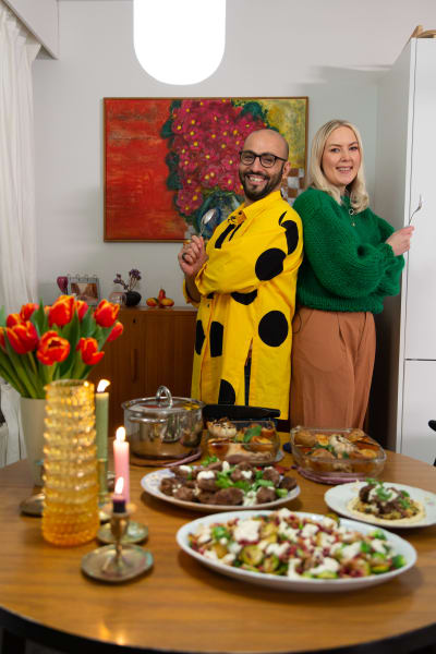 Värikkäästi pukeutunut mies ja nainen poseeraa runsaan ruokapöydän edessä.