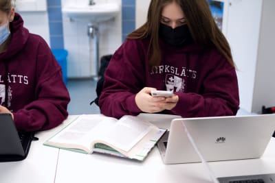 En elev scrollar på sin telefon. Framför sig har hon en dator och en uppslagen bok.