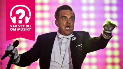 Robbie Williams håller en mikrofon i ena handen och sträcker ut den andra handen framför sig.