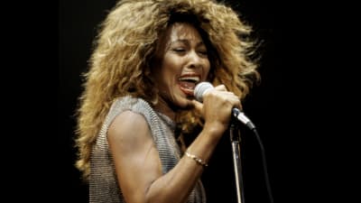 Tina Turner med mikrofon i hand på scen, iklädd en silvrig topp.